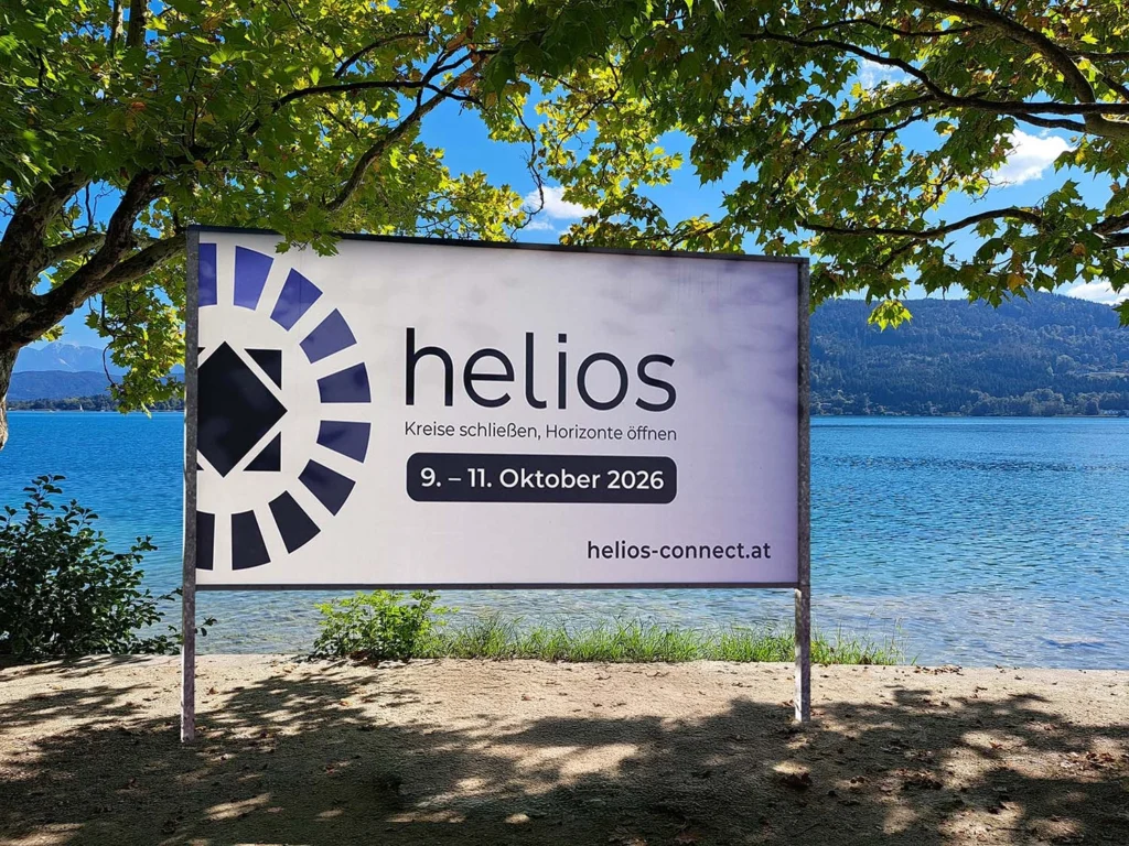 Ein Veranstaltungsplakat vor der Kulisse des blauen Wörthersees. Der Text auf dem Plakat lautet: ‚helios – Kreise schließen, Horizonte öffnen. 9. bis 11. Oktober 2026. helios-connect.at‘. Links neben dem Text befindet sich ein abstraktes, dunkelblaues Logo aus Kreissegmenten.