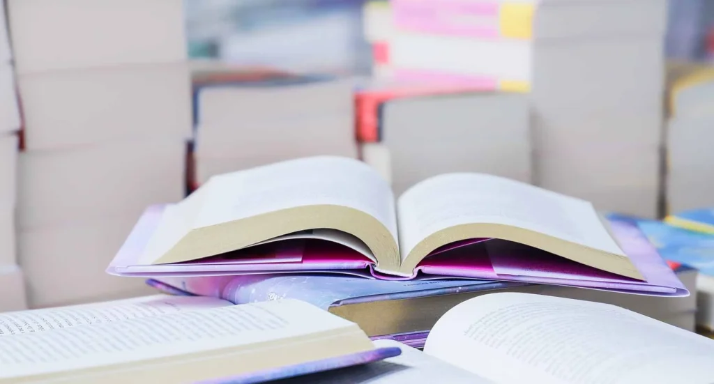 Nahaufnahme eines aufgeschlagenen Buches auf einem Stapel weiterer Bücher in einer hellen, lichtdurchfluteten Umgebung mit unscharfem Hintergrund.